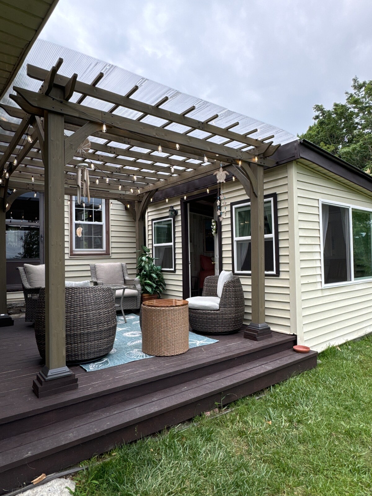 A charming outdoor seating area is showcased under a decorative pergola. Comfortable wicker chairs and a round table are arranged on a wooden deck, which is framed by lush green grass. Subtle string lights hang overhead, adding a soft ambiance to the space.