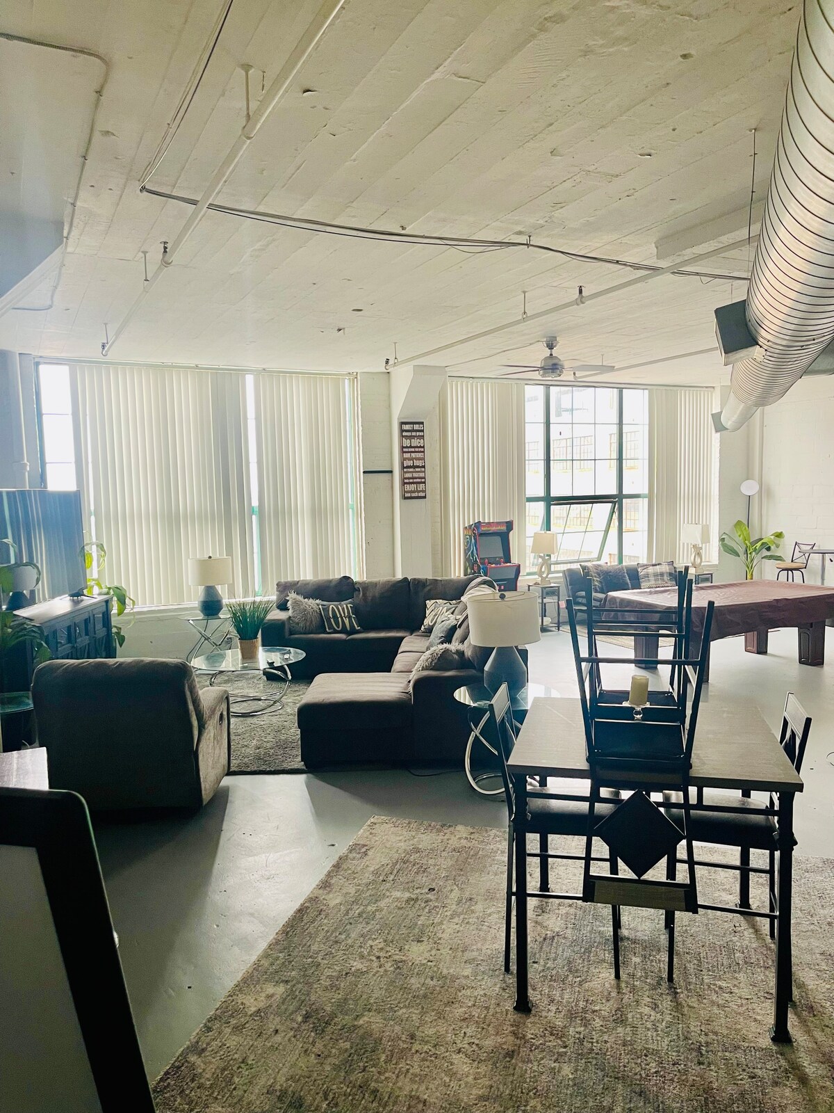 A bright and airy living area features large windows allowing natural light to fill the space. Comfortable seating arrangements include a sectional couch and mix of chairs. A dining table and chairs are positioned nearby, complemented by decorative plants throughout the room.