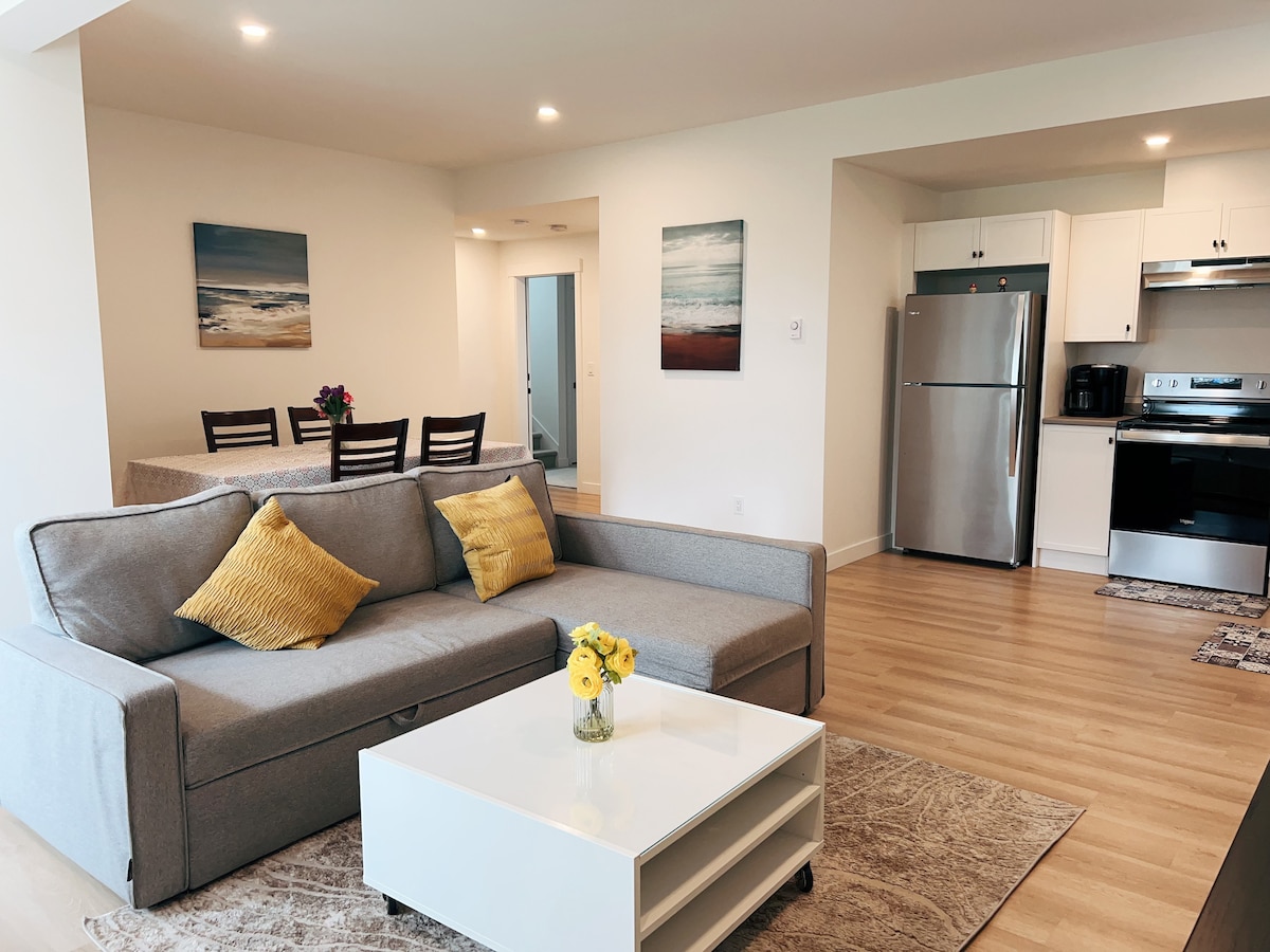 A modern living area displays a light grey sofa adorned with yellow cushions. A white coffee table is centered on a soft area rug. In the background, a dining table with wooden chairs and a fully equipped kitchen with stainless steel appliances can be seen. Artwork on the walls adds a subtle touch.