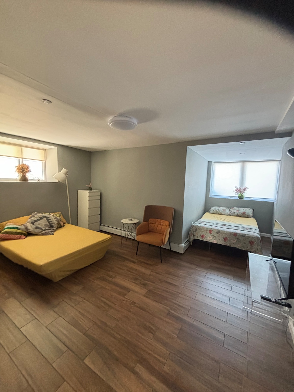 An inviting room features a spacious yellow bed with decorative pillows, complemented by a twin-sized bed adorned with floral bedding. A stylish chair and small side table are positioned nearby, while natural light softly filters through the windows, enhancing the serene atmosphere.