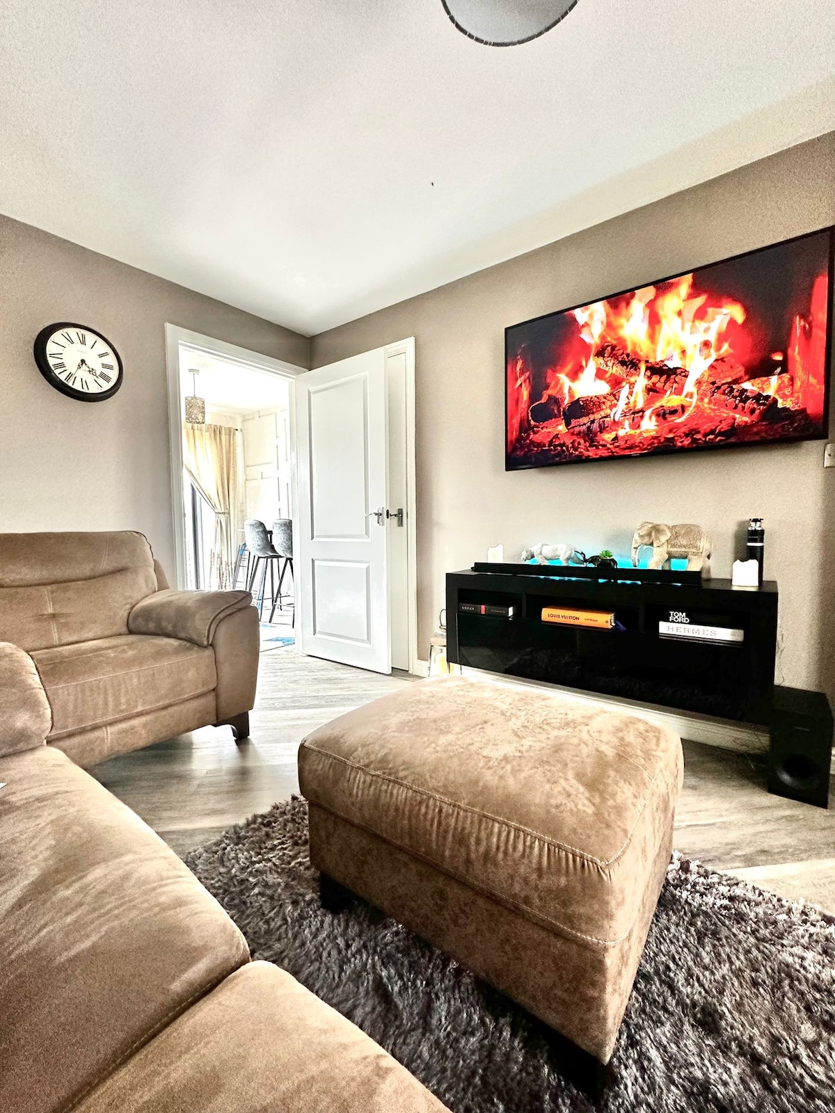 A cozy living area is highlighted by a soft brown couch and an ottoman. A large flat-screen television displaying a fireplace creates a warm ambiance. Light walls contrast with a dark media unit, and a clock on the wall offers a sense of time in this inviting space.