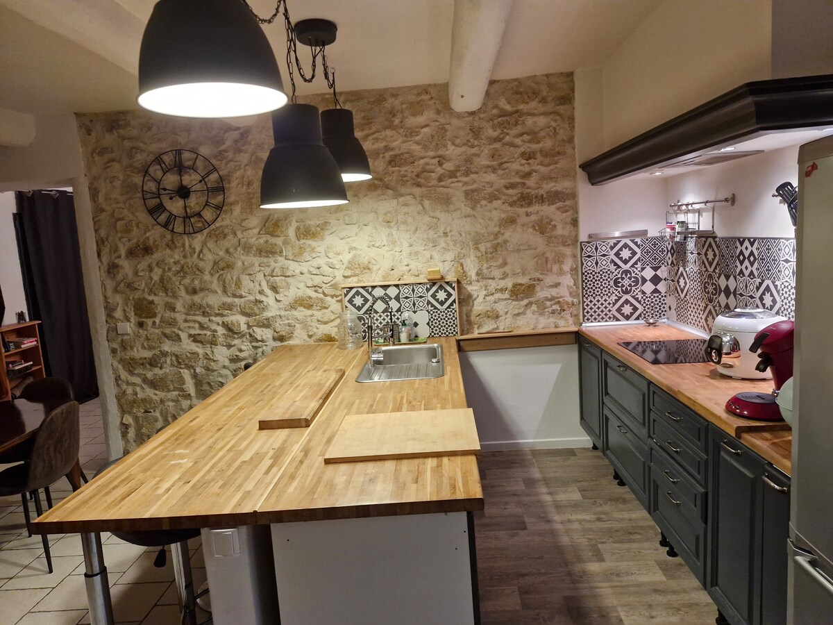 An inviting kitchen space features a wooden countertop and a stone wall, creating a warm atmosphere. Pendant lights hang above the countertop, illuminating the area. A sink is integrated into the countertop, with patterned tiles adding visual interest behind. Kitchen appliances are neatly arranged along the cabinetry.