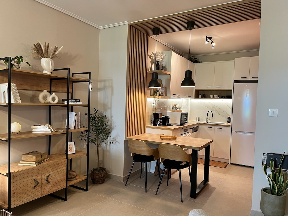 A modern kitchen area is presented with sleek cabinetry and a minimalist design. A wooden dining table with two chairs is positioned next to an open shelving unit displaying decorative items. Soft lighting is provided by black pendant lamps above the table.