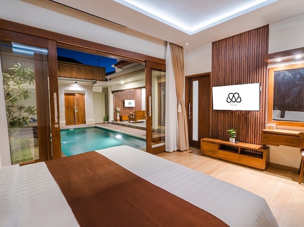 A modern bedroom area features natural wood accents and soft lighting. Large glass doors open to the private pool, offering seamless access to outdoor space. A flat-screen TV and a stylish vanity complete the room, providing a comfortable and functional retreat.