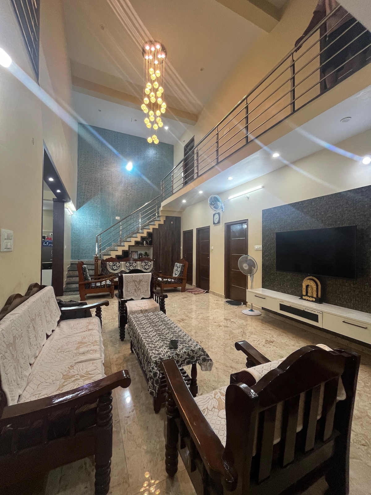 A spacious living area features elegant wooden furniture arranged thoughtfully. A long patterned table sits in the center, surrounded by comfortable seating. The high ceiling hosts a unique chandelier, while a flat-screen TV is mounted on the wall opposite large windows that allow natural light to enter.