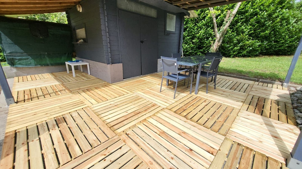 A spacious outdoor deck features alternating patterns of wooden planks, providing ample space for relaxation. A simple table with four chairs is positioned for dining. The area is surrounded by greenery, contributing to a calm and open environment.