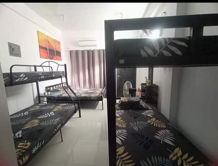 The room features two bunk beds with floral-patterned bedding. A fan is placed nearby, and a window with light-filtering curtains allows natural light to enter. A small storage unit and some personal items can be seen in the background.