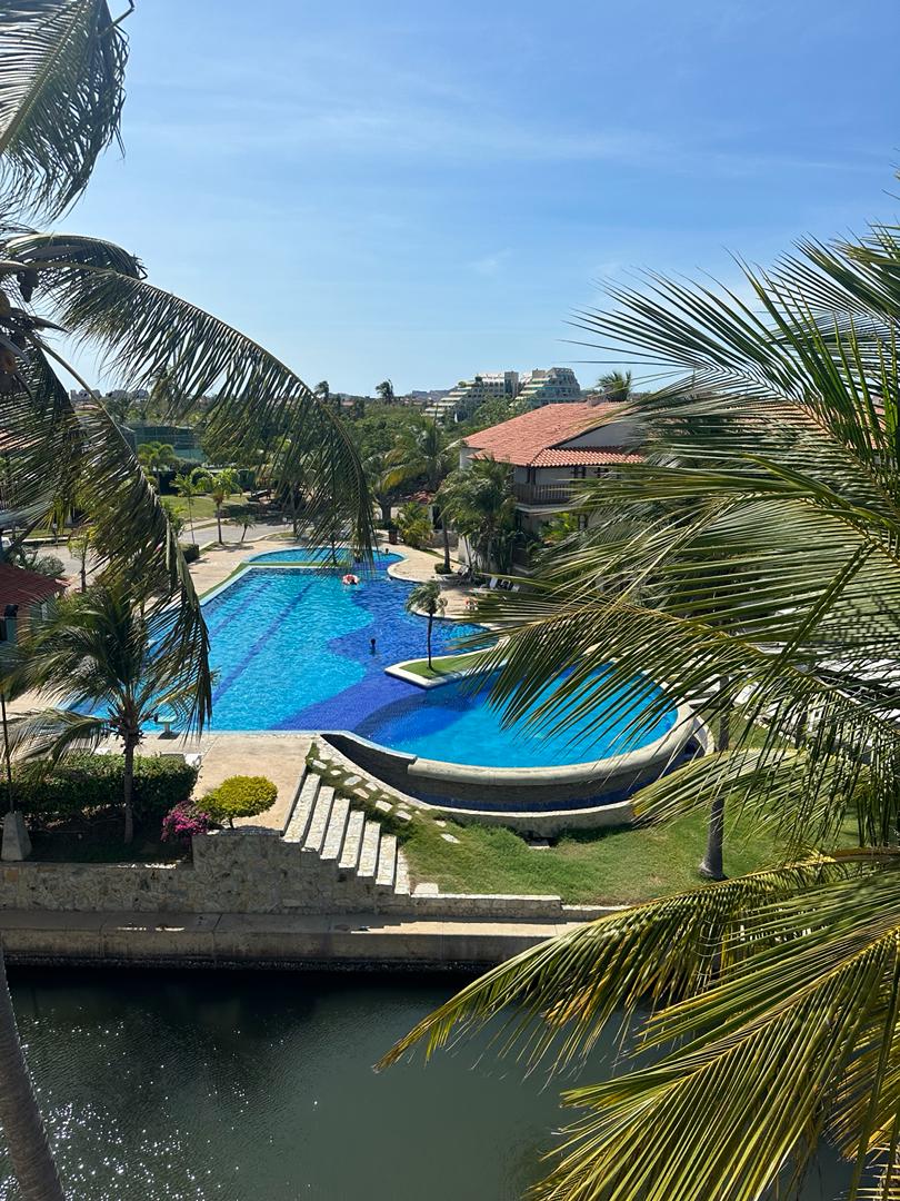 An expansive swimming pool is surrounded by lush greenery and palm trees. Its bright blue waters reflect the clear sky, while lounge areas are arranged along the edges. The scene is complemented by a canal flowing nearby, enhancing the serene atmosphere.