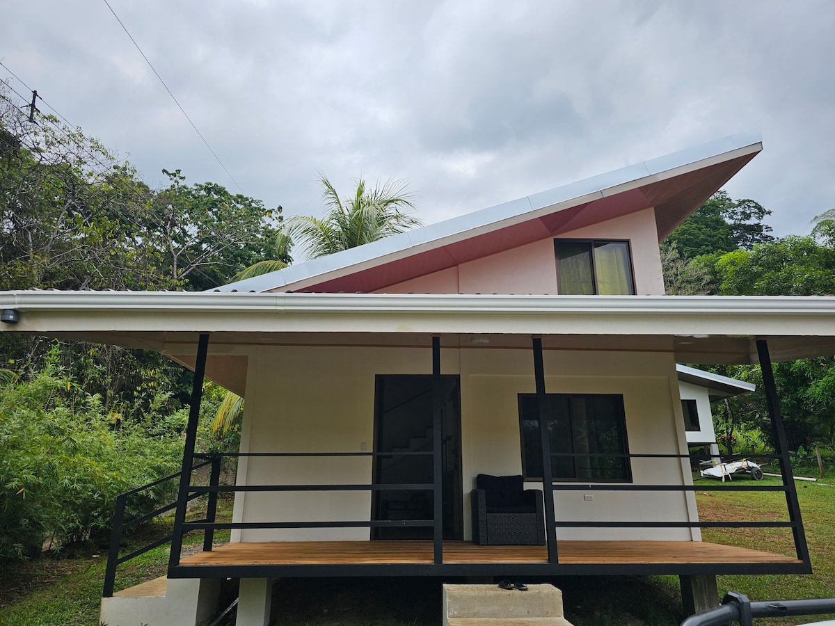 A modern house features a unique sloping roof and a covered porch area. Neatly arranged outdoor furniture is present on the wooden deck, while a large window allows natural light into the living space. Lush greenery surrounds the property, creating a serene environment.