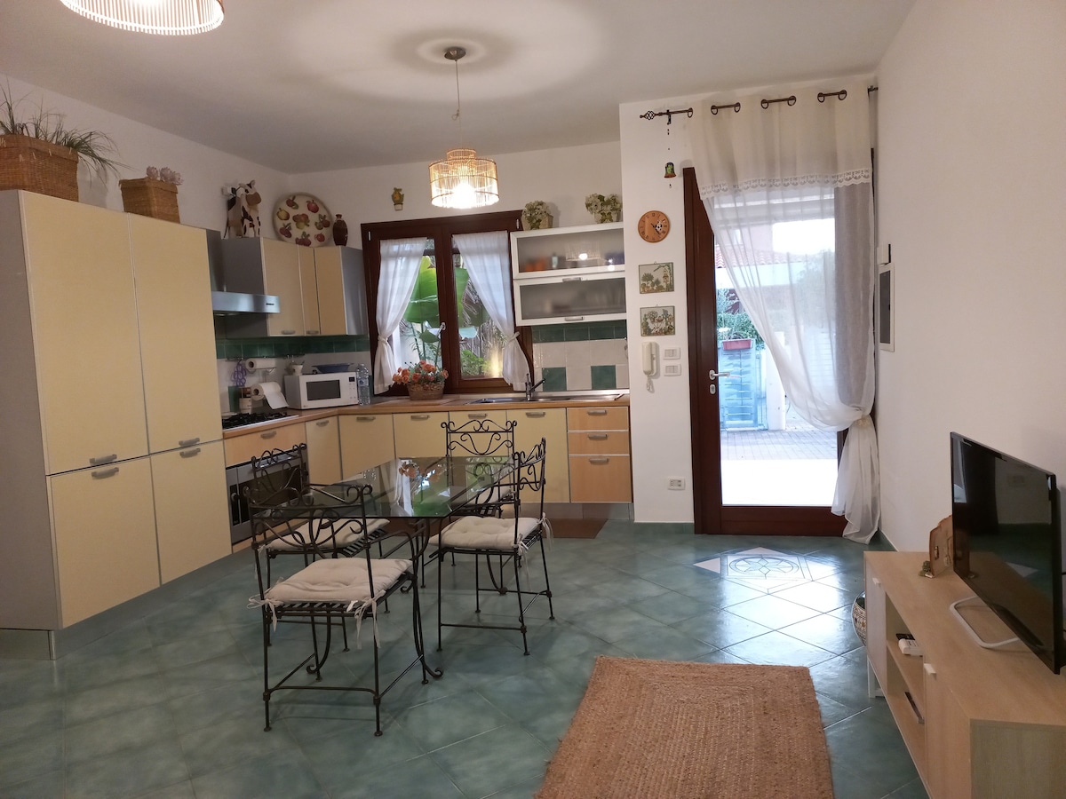A bright kitchen area features modern cabinets in soft yellow tones, complemented by a glass-top dining table surrounded by four iron chairs. Natural light enters through large windows adorned with sheer curtains, creating an inviting space suitable for meals and gatherings.