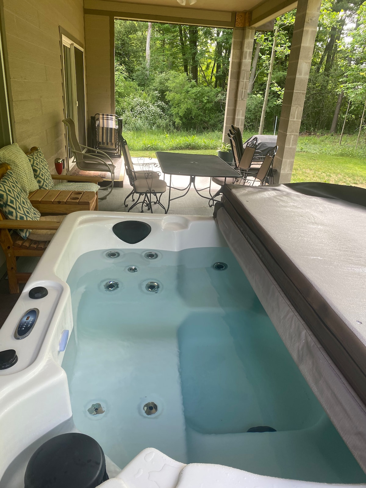 A hot tub is situated on a covered porch, featuring multiple jets and a secure cover. Nearby, a dining table with four chairs offers a relaxed outdoor setting, surrounded by lush greenery. Comfortable seating options add to the tranquil atmosphere.