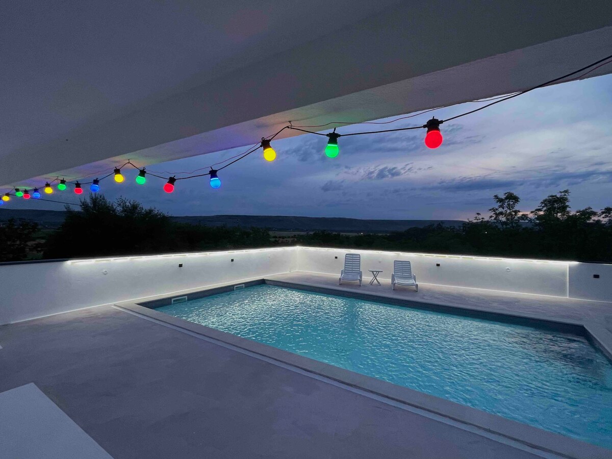 An outdoor pool area is featured, illuminated by colorful lights strung above. Two loungers are positioned beside the sparkling water, which reflects the evening sky. The surrounding space offers a serene view of the landscape, creating a peaceful environment for relaxation.