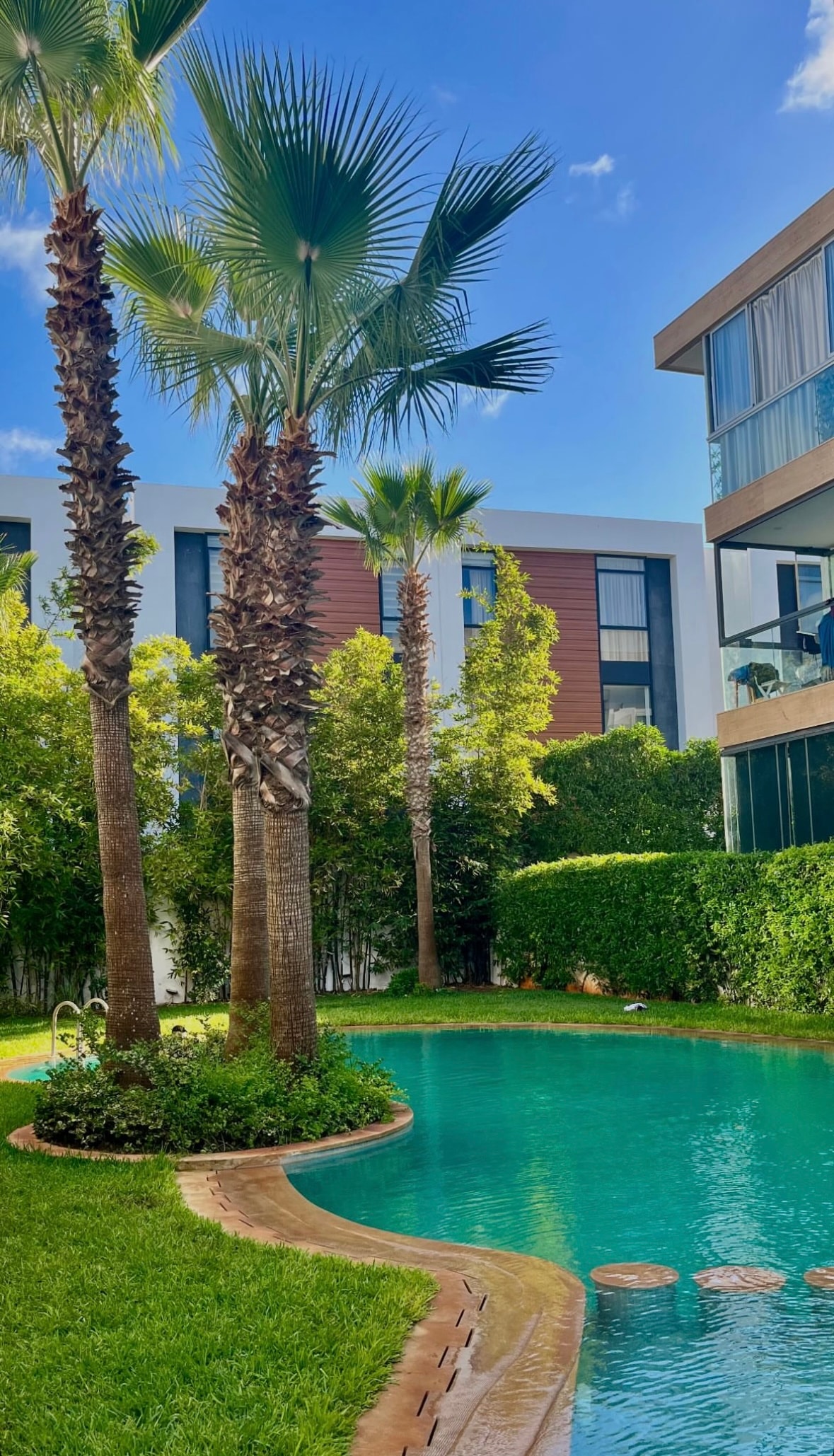 A tranquil outdoor area features a curved swimming pool surrounded by lush green grass and palm trees. The modern building façade is visible in the background, with wooden elements contrasting against the greenery. Bright blue skies provide a serene atmosphere.