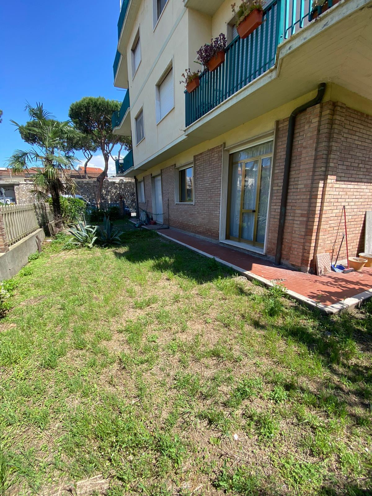 A spacious garden area is visible, featuring lush green grass and bordered by light-colored brickwork. The entrance to the home is framed by glass doors, with planters adorned with flowers on the balcony above. Surrounding trees and plants contribute to a tranquil outdoor setting.