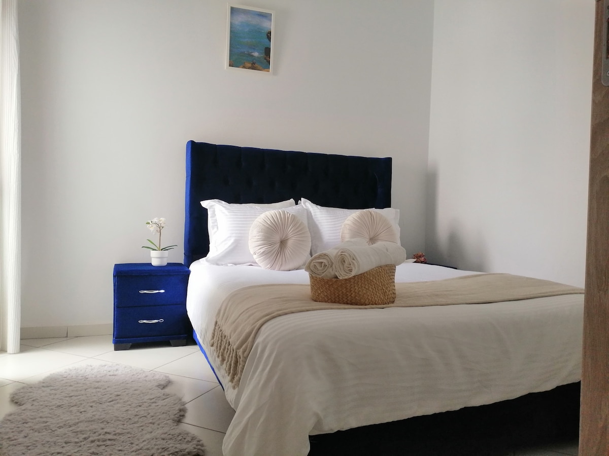 A restful sleeping area is featured, showcasing a spacious bed with a tufted dark blue headboard and neutral-toned bedding. A bedside table is positioned beside the bed, providing a lamp and decorative plant. Soft textures are added with a round rug beneath the bed.