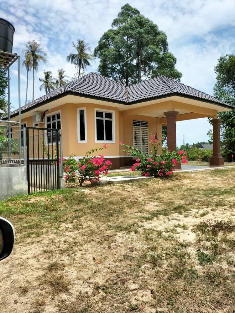 A single-story house is framed by lush greenery and blooming pink flowers. The exterior features a textured orange finish with a dark tiled roof. A covered porch is supported by two elegant columns, while a gated entrance leads to the property.
