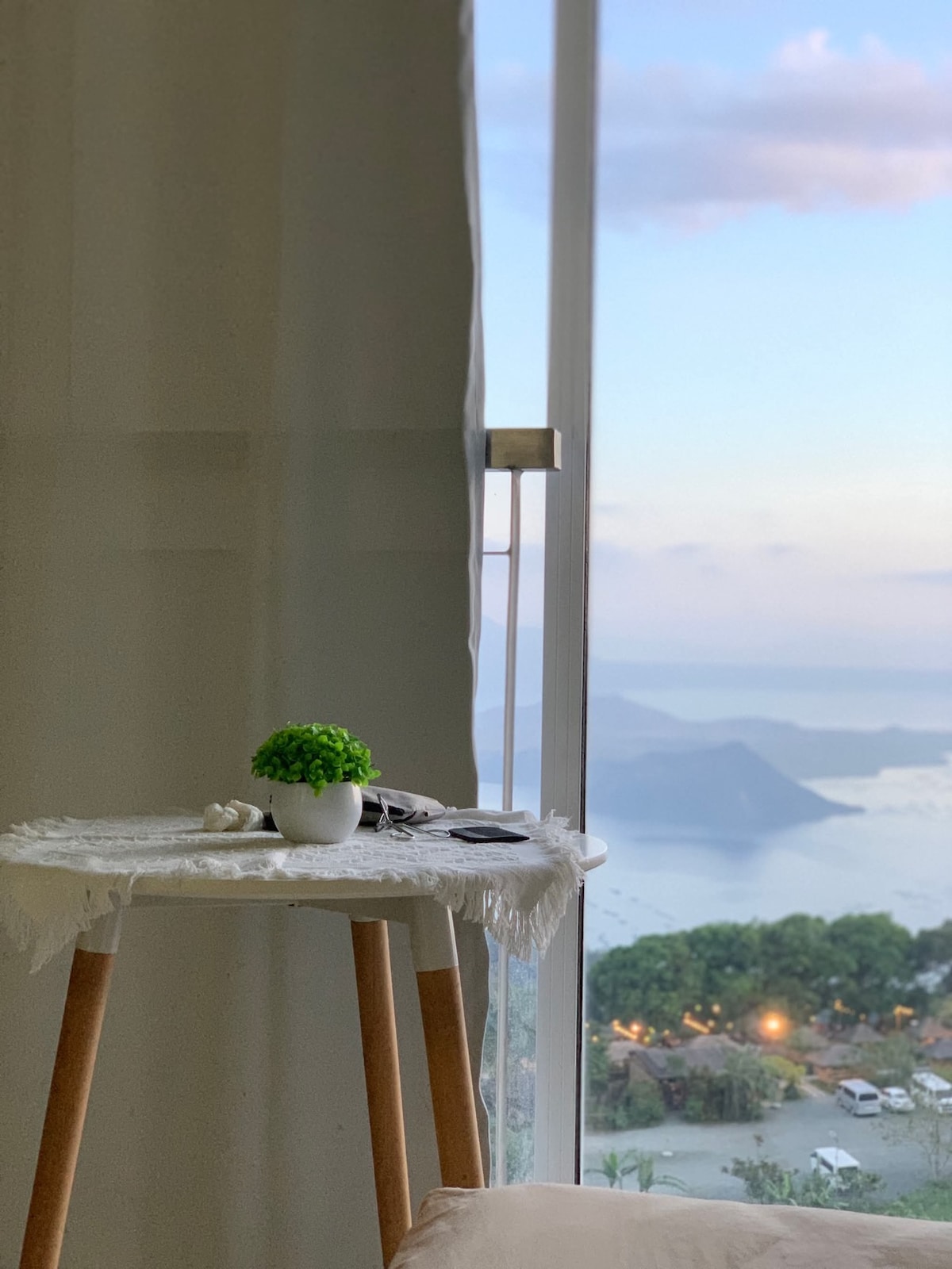 A small round table with wooden legs is positioned next to a large window, offering a scenic view of the Taal Lake and surrounding landscape. A potted plant adorns the table, and personal items are neatly arranged beside it.