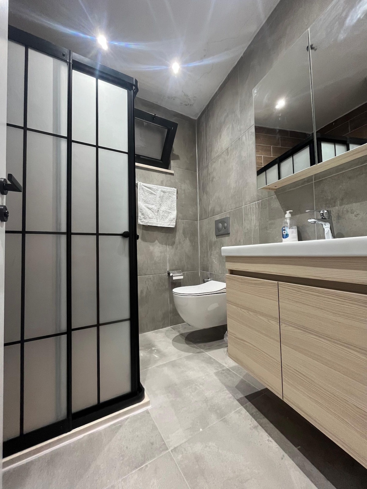 A modern bathroom is presented, featuring a sleek, enclosed shower space with frosted glass panels. The toilet is positioned adjacent to a contemporary wooden vanity, complete with a white countertop. Natural light filters through the small window, illuminating the neutral-colored tiles.