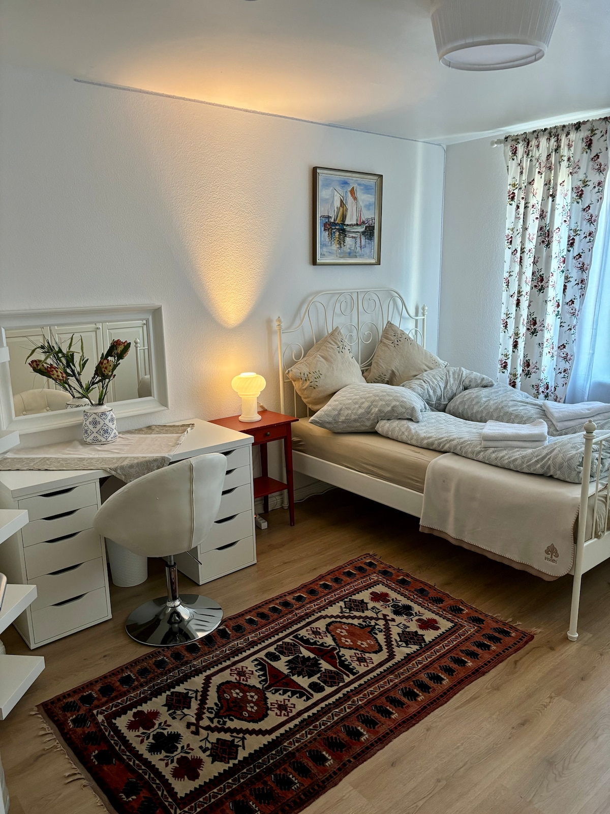 A bright bedroom features a spacious bed adorned with light bedding. A desk with a chair is positioned beside the bed, accompanied by a bedside table holding a lamp. A patterned rug lies on the floor, complementing the floral curtains and artwork on the wall.