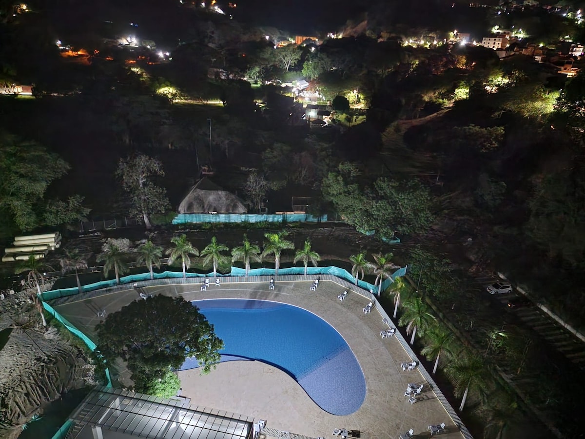 An outdoor swimming pool is framed by lush palm trees, with several lounge chairs arranged around its edges. The area is subtly illuminated, revealing a distant landscape of hills and buildings under a night sky.