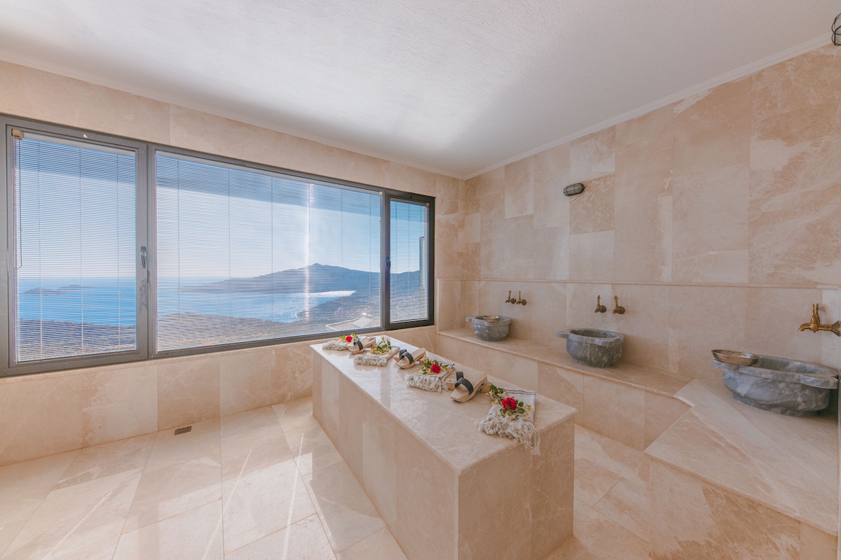 A bright and serene bathroom features a large countertop made of stone, accompanied by two sinks. Expansive windows frame a panoramic view of the sea and surrounding landscape, enhancing the tranquil environment. Natural light illuminates the polished stone flooring, creating a calm atmosphere.