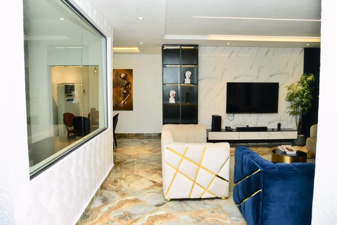 A stylish lounge area is presented, featuring comfortable seating in neutral and blue tones. A large flat-screen television is mounted on the wall, with decorative shelves displaying art pieces. The floor is adorned with a marbled design, complementing the modern aesthetic of the space.