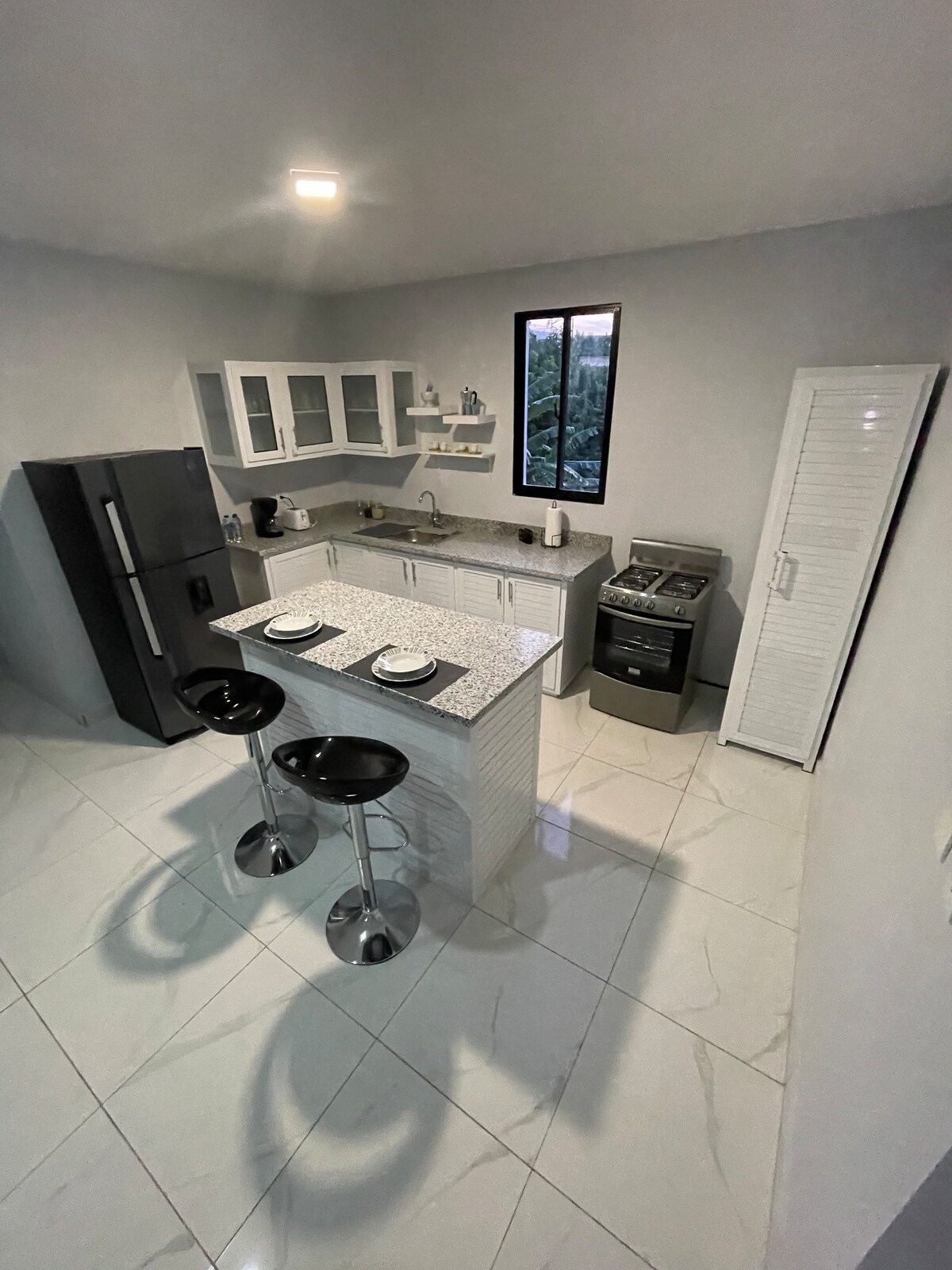 A modern kitchen features sleek cabinetry with glass-fronted upper shelves. Two bar stools are positioned at a stylish countertop. Stainless steel appliances, including a refrigerator and oven, complement the light-colored tiled floor, while a window allows natural light to illuminate the space.