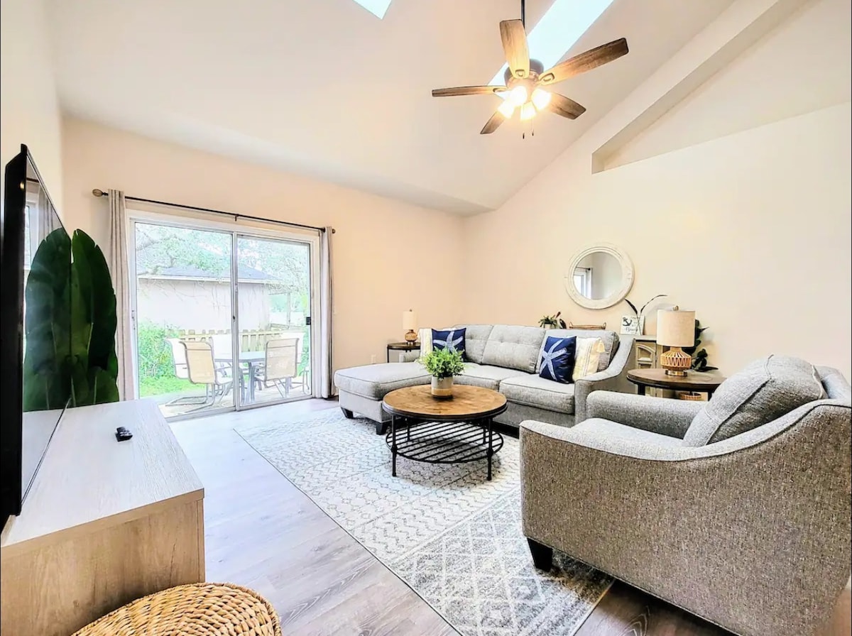 A bright living area features a comfortable sectional sofa and an accent chair, set against a backdrop of vaulted ceilings and natural light pouring in from the skylights. A circular coffee table rests on a patterned rug, with sliding doors leading to an outdoor seating area.