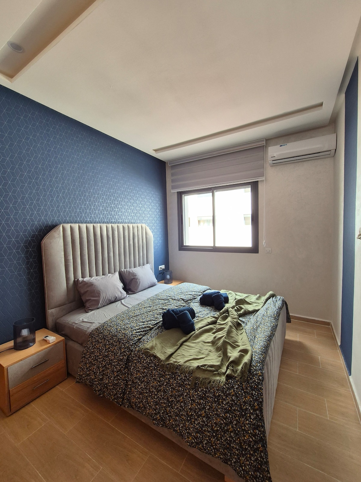 A neatly arranged bedroom features a spacious bed dressed in a textured blanket and subtle patterned cover. Two gray pillows rest at the headboard, beside a bedside table with a lamp. Natural light enters through a window dressed with blinds, enhancing the clean, contemporary design.