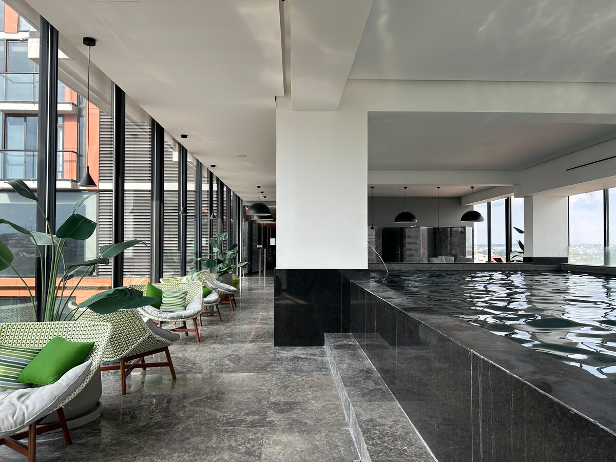 A modern indoor pool area features large windows, allowing natural light to fill the space. Comfortable seating with green accents is positioned nearby, complemented by decorative potted plants. The pool reflects the surrounding views, creating a serene atmosphere for relaxation.