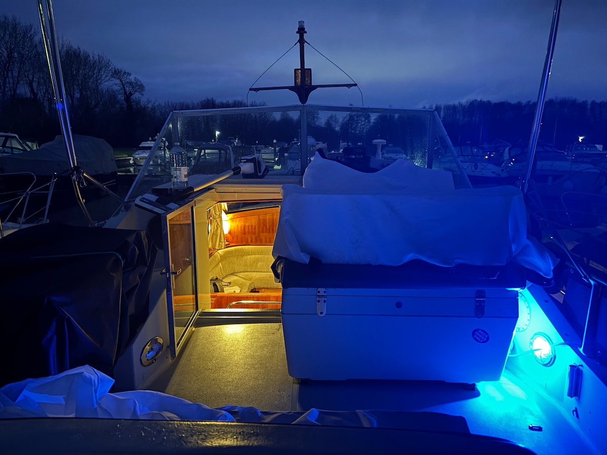A serene evening scene on the boat is depicted, featuring soft blue ambient lighting. An inviting space is shown with a neatly covered seating area and a nearby door that leads to the interior, creating a cozy atmosphere. The tranquil waterways are suggested in the background.
