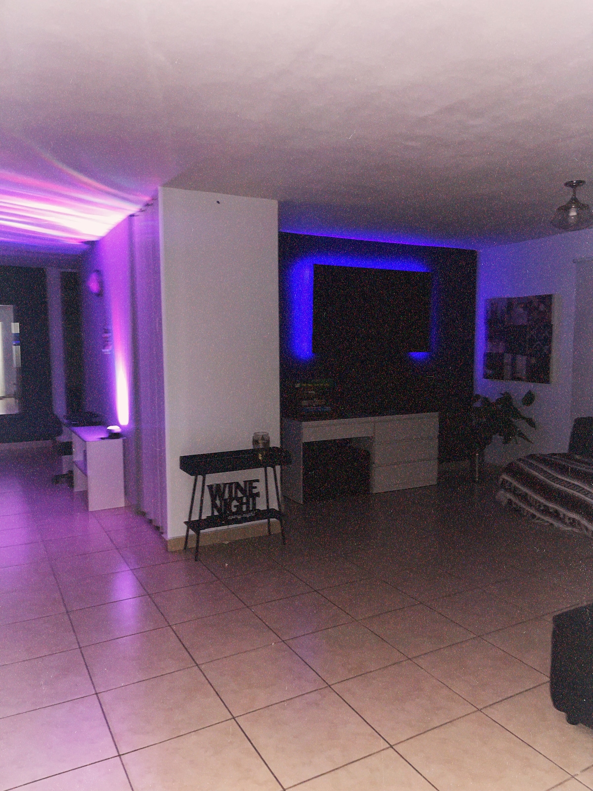 A spacious living area is illuminated by subtle purple lighting, creating an inviting ambiance. A black sofa is positioned against the wall, complemented by a striped blanket. A decorative table with a 'WINE' sign is present, alongside a sleek entertainment unit.
