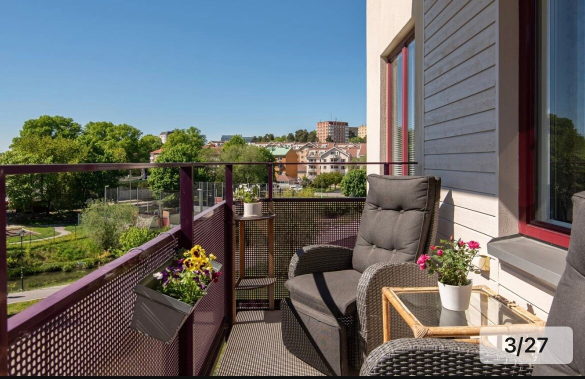 A sunny balcony features two comfortable chairs with soft cushions, accompanied by a small wooden table. Flower boxes are filled with vibrant blooms, providing a touch of color. The surrounding view includes greenery and buildings, enhancing the relaxing outdoor atmosphere.
