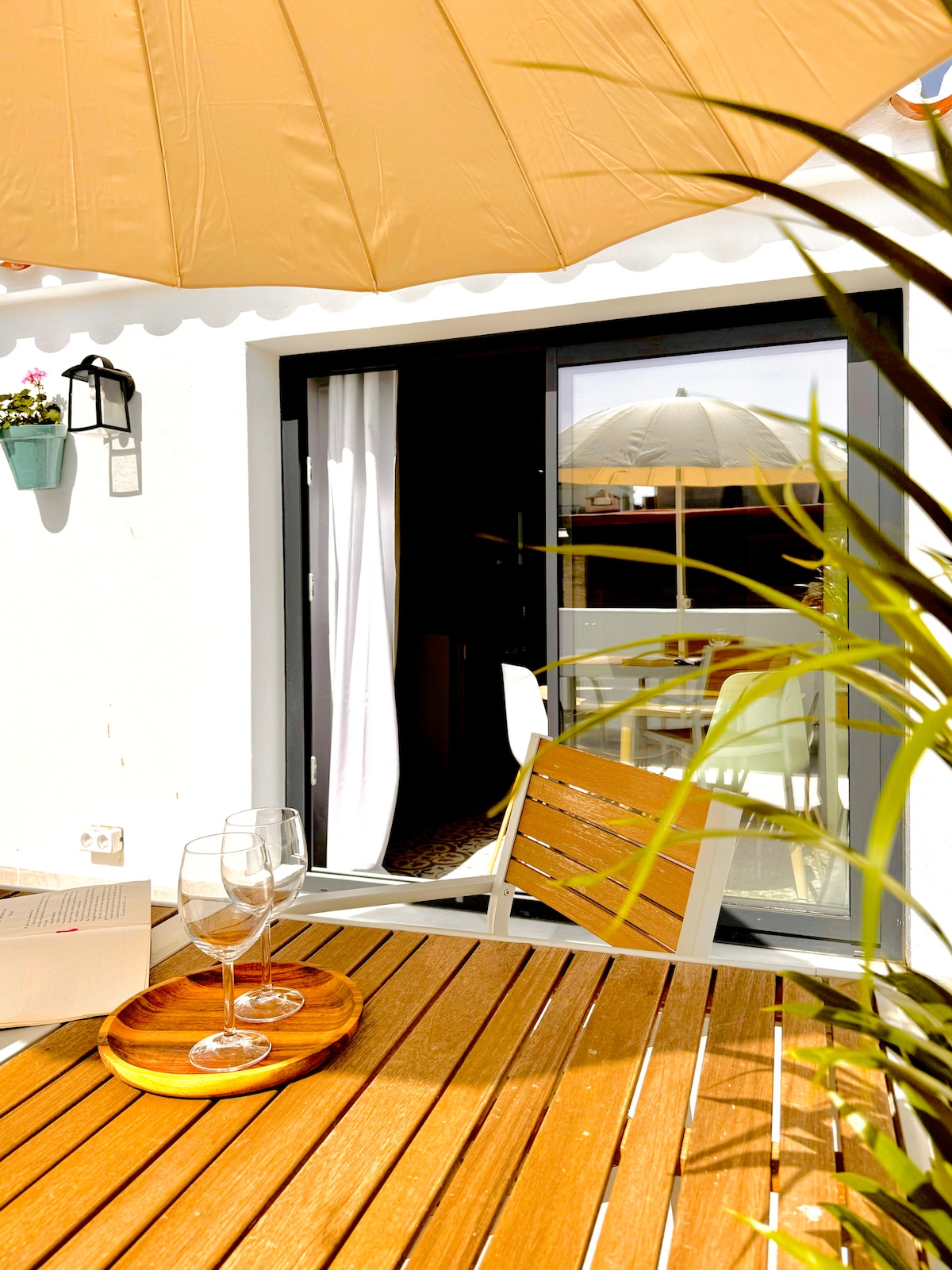 An outdoor terrace is presented with a wooden dining table and matching chairs under a sun umbrella. Glasses and a wooden tray are placed invitingly on the table. A sliding glass door leads into the interior space, framed by minimal greenery.