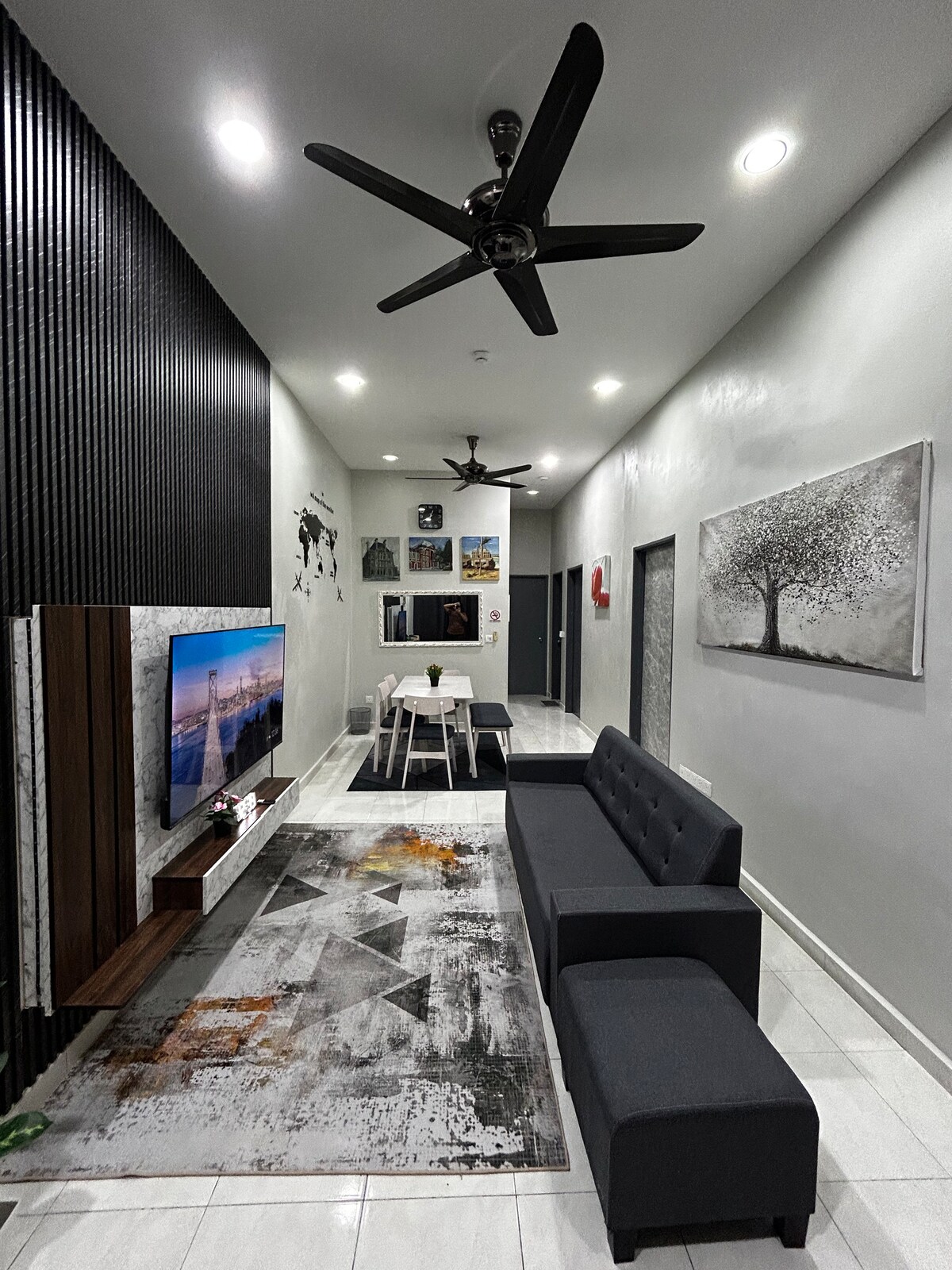 A modern living area is depicted with a sleek gray couch and an entertainment unit featuring a flat-screen TV. Stylish artwork adorns the walls, and a dining table for four is positioned nearby. Ceiling fans provide a refreshing touch, and an area rug adds warmth.