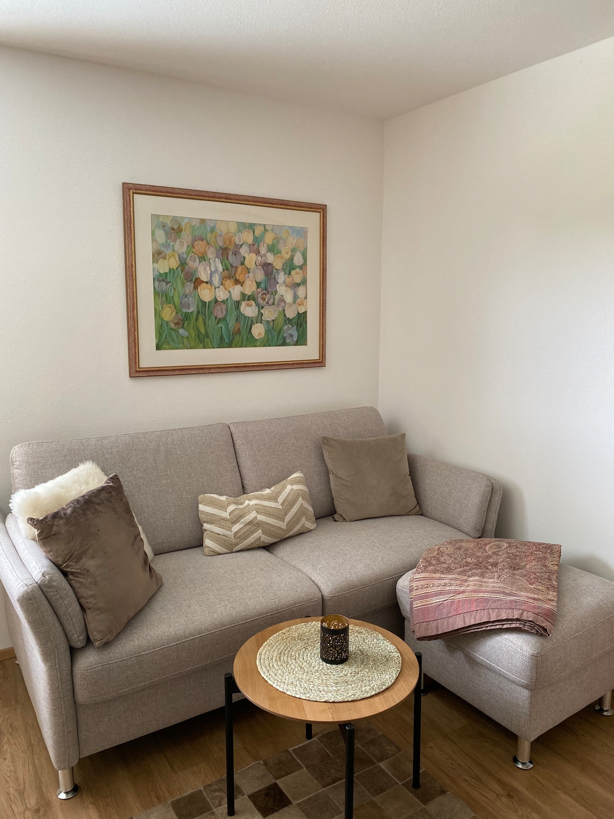 A light-filled corner of the living area features a comfortable sofa with decorative cushions. A round wooden table with a dark centerpiece is positioned nearby. A colorful floral painting adorns the wall, complementing the neutral tones of the furniture.