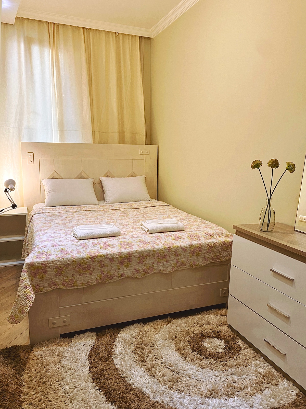 A serene sleeping area is presented with a spacious bed adorned with a light floral bedspread. Two folded towels are neatly placed on the bed. Natural light streams in through sheer curtains, illuminating the room. A simple dresser is positioned beside a mirror.