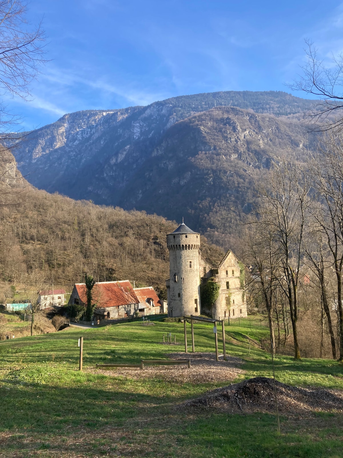 A historical castle is set against a backdrop of steep mountains, surrounded by lush greenery. The stone structure features a tower and arched windows, while the nearby buildings are characterized by traditional red-tiled roofs. The area offers open grassy spaces for relaxation.