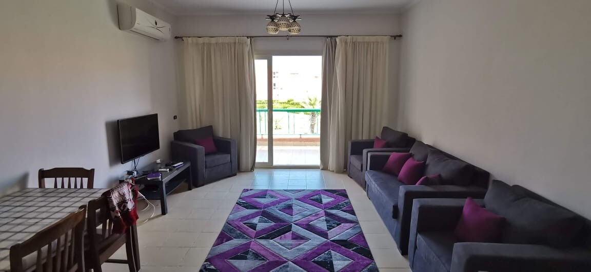 A spacious living room is furnished with a dark sofa set, paired with accent cushions. A dining table is positioned nearby. Large glass doors open to a balcony, surrounded by curtains. A geometric patterned rug covers the floor, offering a touch of colour.