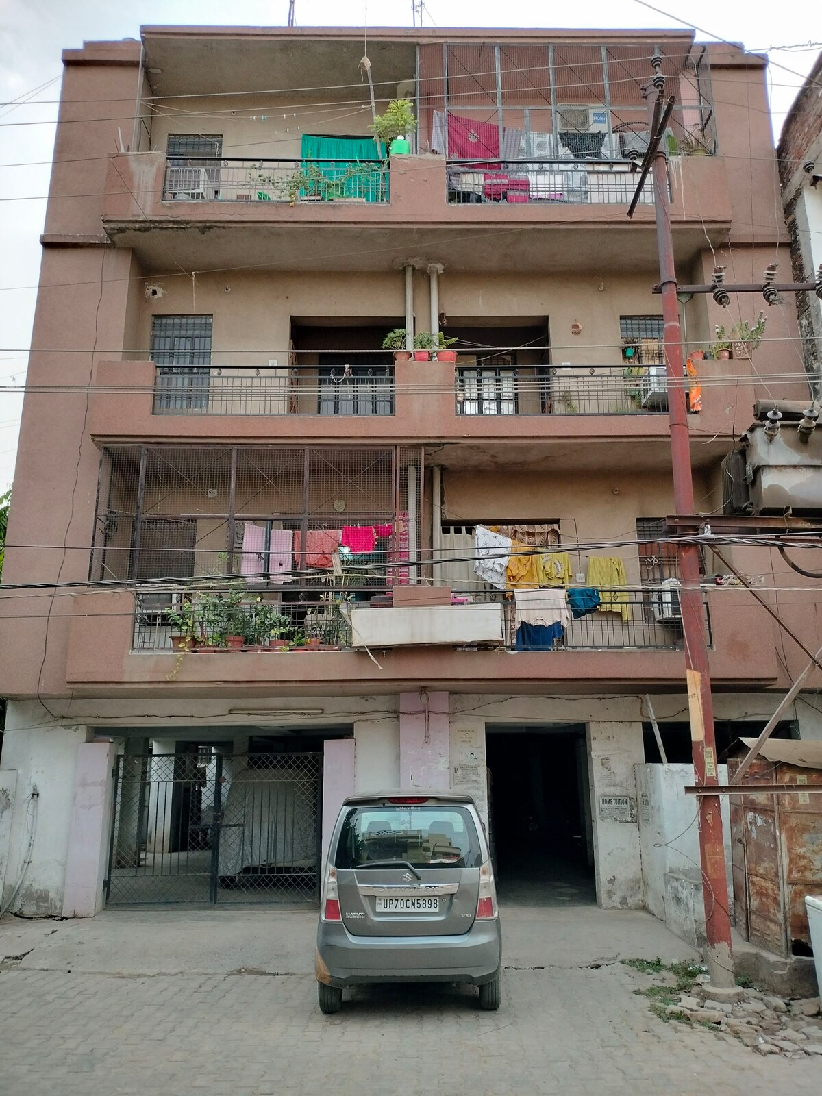 A three-story building is depicted with a light brown facade. Balconies on each floor are adorned with various plants and colorful clothes hanging to dry. A parked car is visible in the foreground, leading to the entrance of the building.