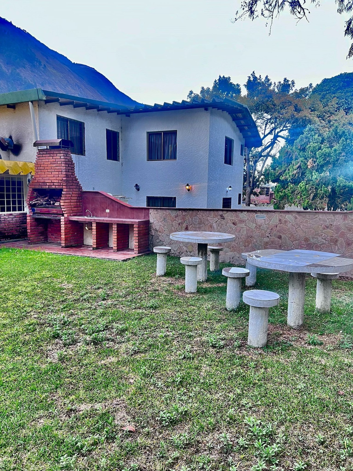 An outdoor space features a stone table surrounded by several stools, set on a lush green lawn. A brick barbecue area is visible nearby, accompanied by a stone cooking surface. The background includes a white chalet and mountainous scenery.