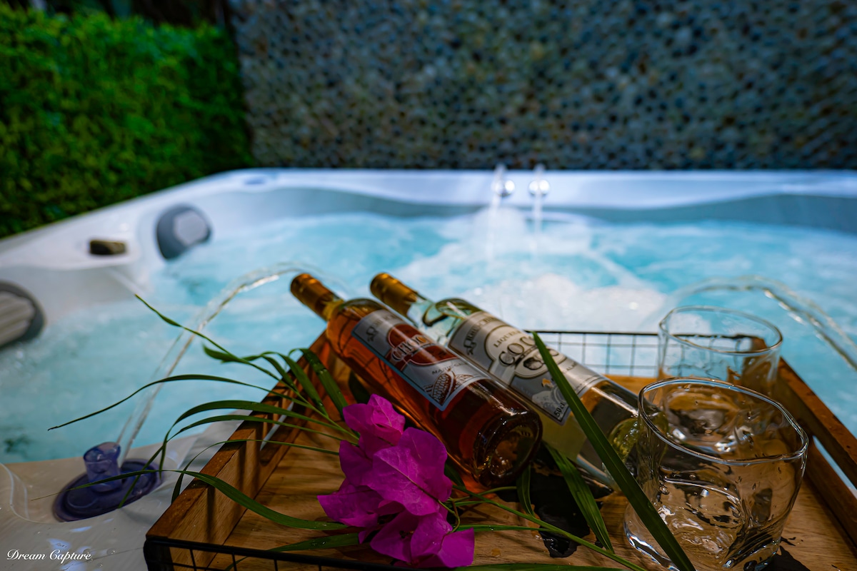 A jacuzzi with bubbling water is surrounded by lush greenery. A wooden tray holds three bottles of wine and two glass cups, complemented by a vibrant pink flower, creating a serene setting for relaxation.
