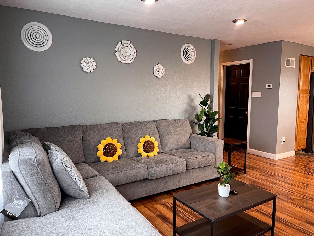 A comfortable living area is depicted with a gray sectional sofa, accented by sunflower pillows. A small wooden coffee table sits in front, accompanied by a potted plant. Decorative wall art adds a subtle touch to the neutral gray backdrop, creating a welcoming space.