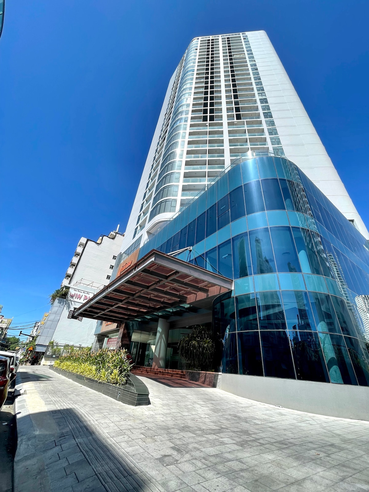 A modern high-rise building is depicted, featuring a striking facade of blue-tinted glass and clean lines. The entrance is highlighted by a spacious overhang and landscaped greenery, offering a welcoming atmosphere. The clear blue sky creates a bright backdrop for the architectural design.