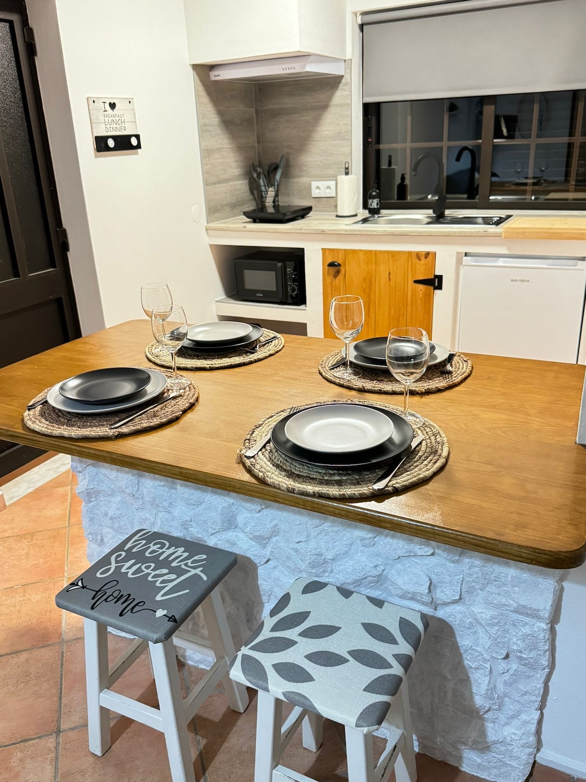 A cozy dining area features a wooden table set for four, with circular placemats, plates, and wine glasses. Nearby, a small kitchen area is visible, equipped with a microwave and a sleek countertop, complemented by warm, neutral tones throughout.