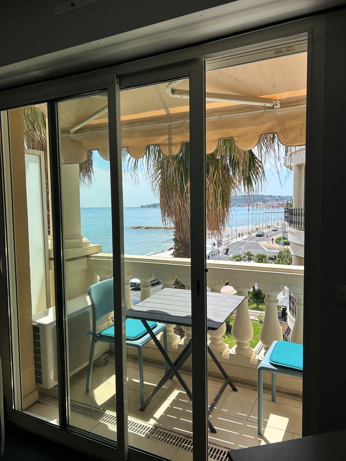 A balcony view showcases a serene seascape, with palm trees framing the scene. A small table with two blue chairs is placed against the railing, allowing for relaxation while overlooking the water. The architecture features large glass doors that invite natural light indoors.