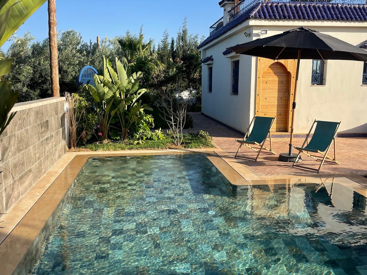 A clear swimming pool reflects the blue sky, surrounded by a landscaped area with greenery. Two lounge chairs are positioned under a large umbrella, offering a shaded retreat. The house features a wooden door and a tiled roof, contributing to a peaceful outdoor atmosphere.