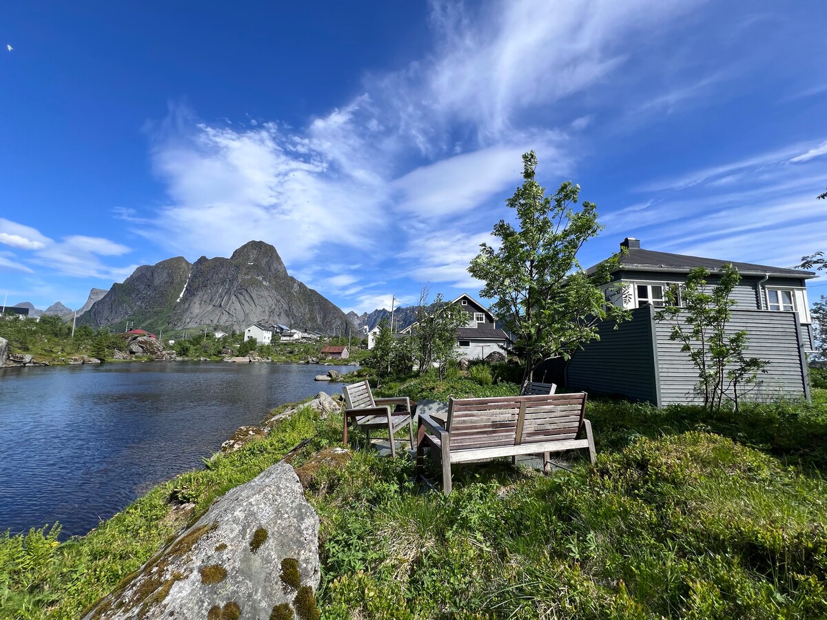 A serene outdoor area features two wooden benches positioned by a tranquil body of water. Behind them, a scenic view of towering mountains and quaint houses can be seen under a blue sky adorned with scattered clouds.