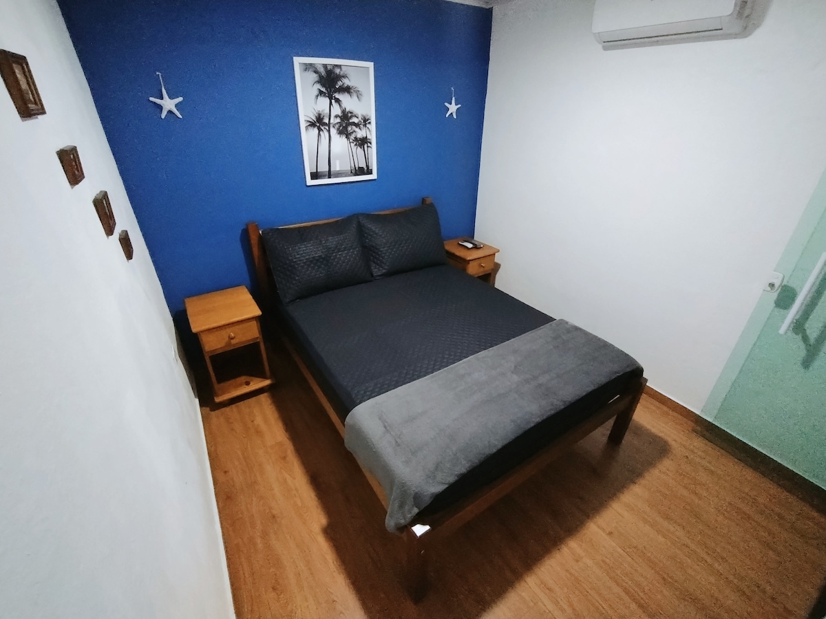 A cozy bedroom is presented with a spacious bed covered in dark bedding. A blue accent wall features a framed image of palm trees. Two wooden nightstands flank the bed, and a white ceiling and air conditioning unit are visible.