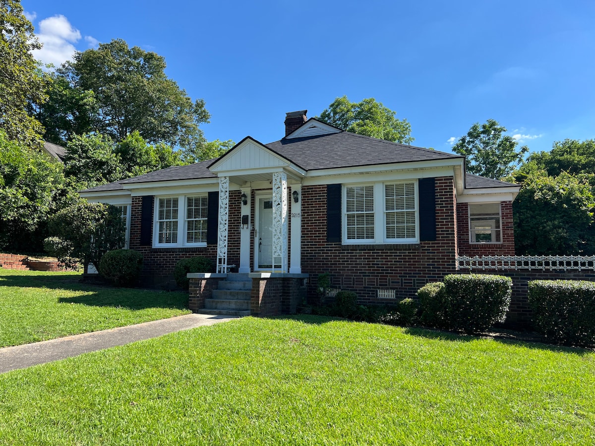 A single-story brick bungalow is positioned on a manicured lawn under a clear blue sky. White columns support a front porch, with large windows flanking the entrance. Lush greenery surrounds the property, creating a serene and inviting exterior.